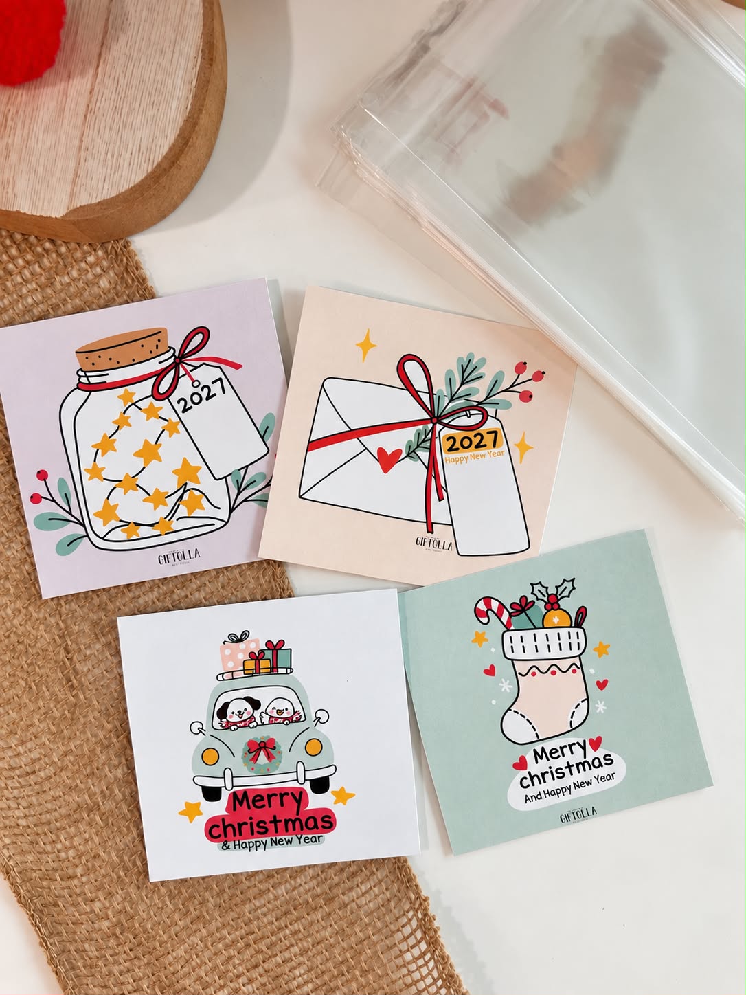 Close-up of four illustrated Christmas and New Year cards beside clear bags on burlap