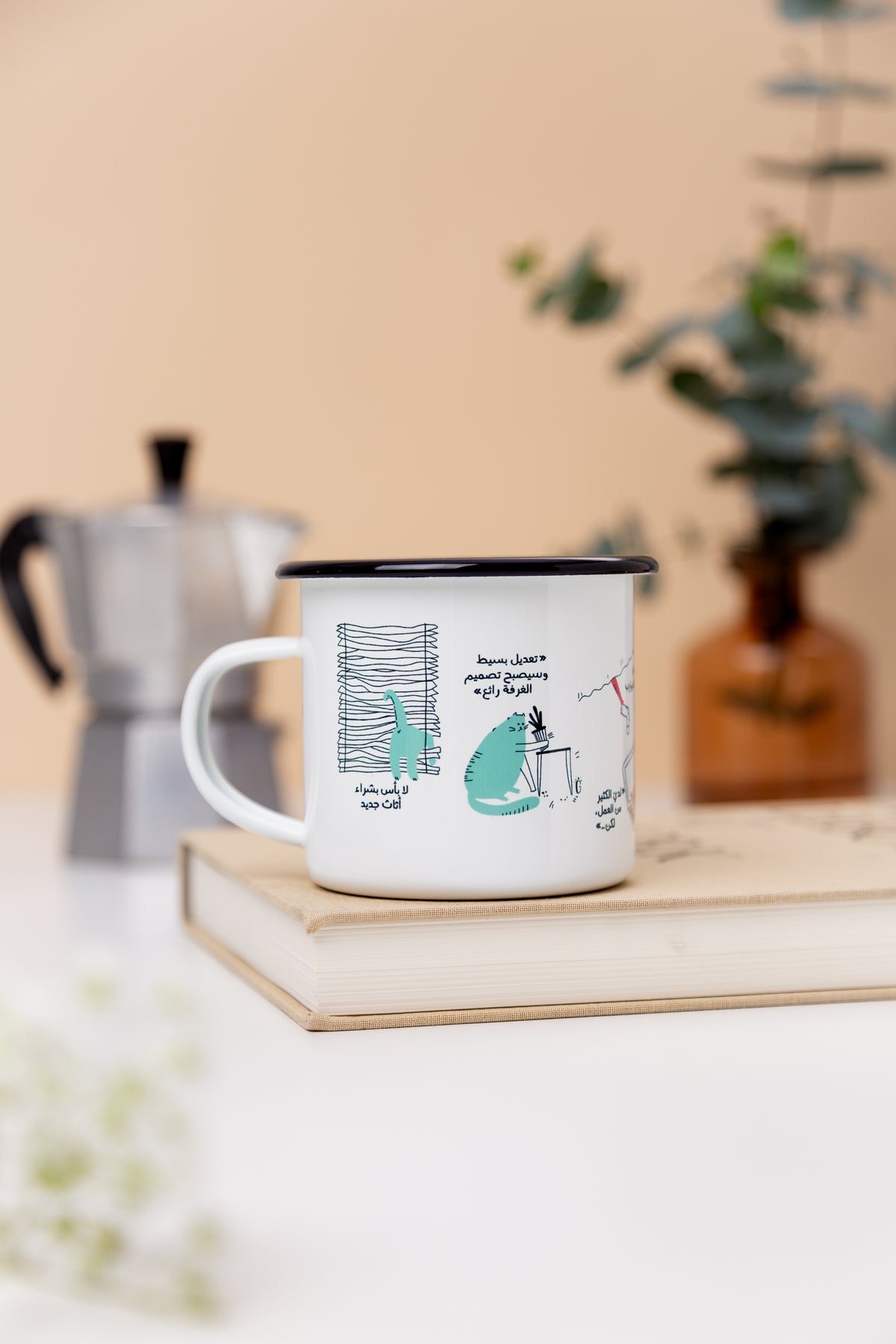 White enamel mug with a teal cat illustration on a book beside a moka pot, Cat enamel mug by Giftolla