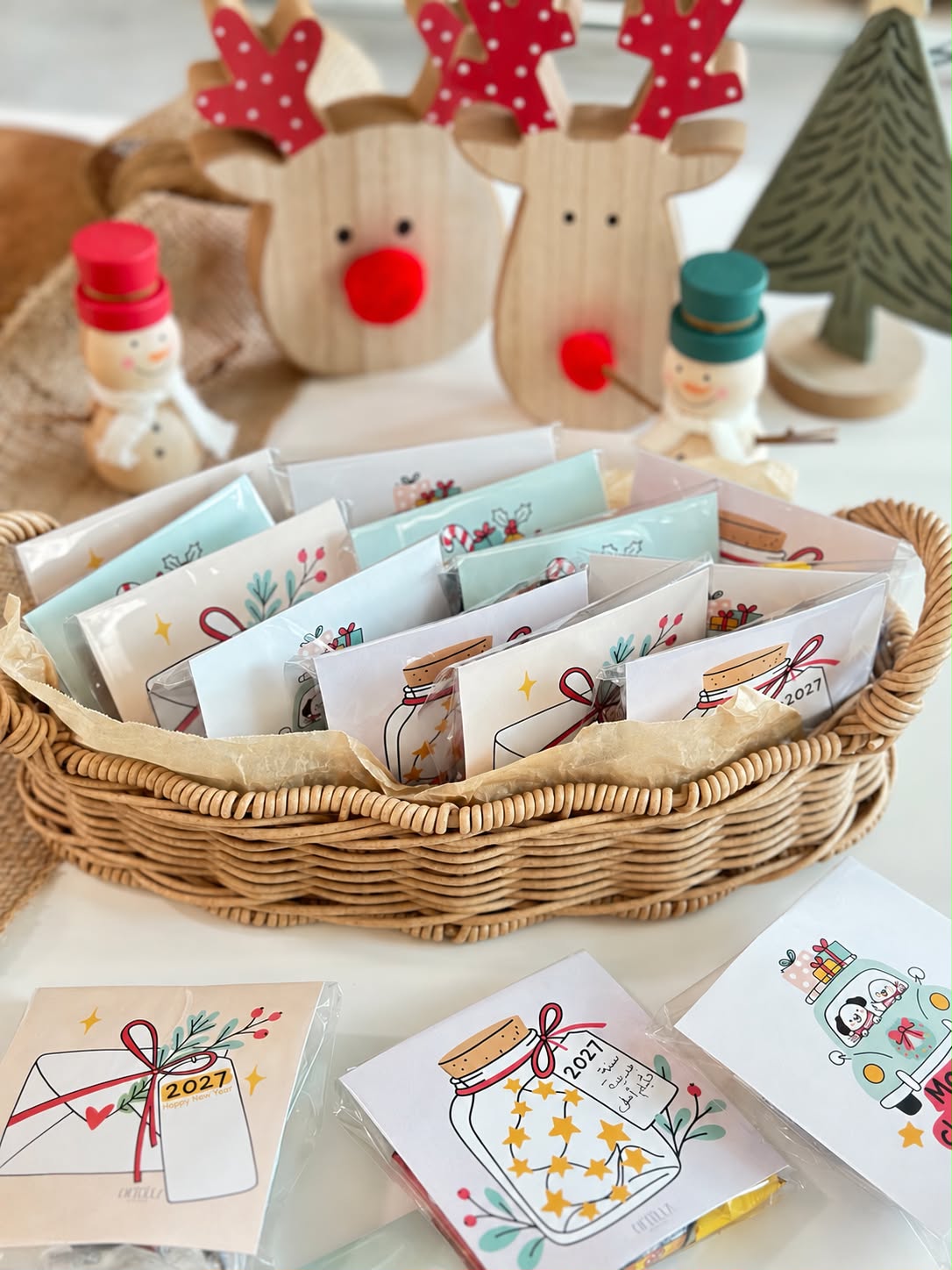 Wicker basket filled with Christmas cards packed in clear bags, styled with wooden reindeer and snowmen