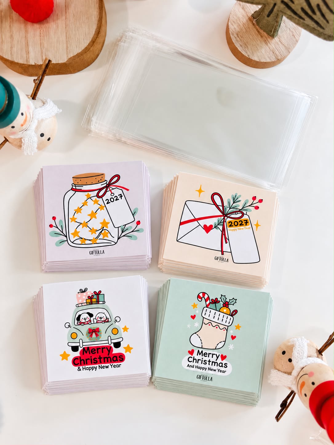 Four stacks of illustrated Christmas and New Year cards with a stack of clear bags beside wooden reindeer decor